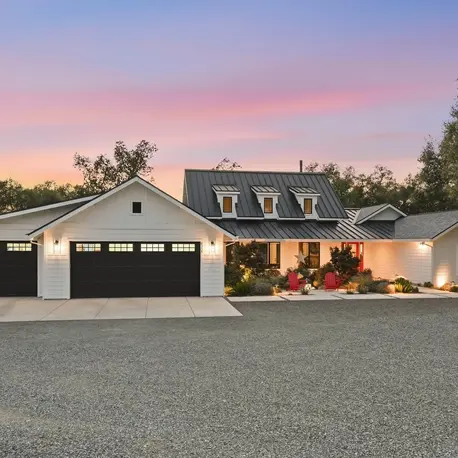 Front elevation at dusk with three-car garage wing, dormers, and lit landscaping