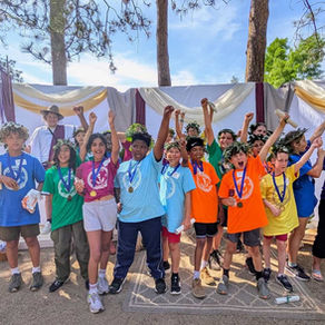 Smiling kids in colorful shirts and laurel crowns cheer with raised arms on an outdoor stage under trees and draped fabric.