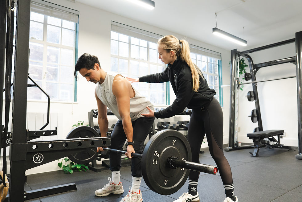 A woman assists a man in a gym doing a barbell deadlift. Bright room with large windows and gym equipment. Focused and instructional mood.