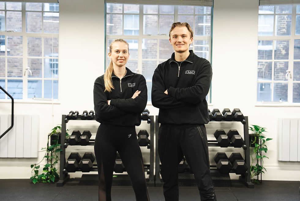 Two people in black uniforms stand with arms crossed in a gym, smiling. Background shows windows, dumbbells, and plants.
