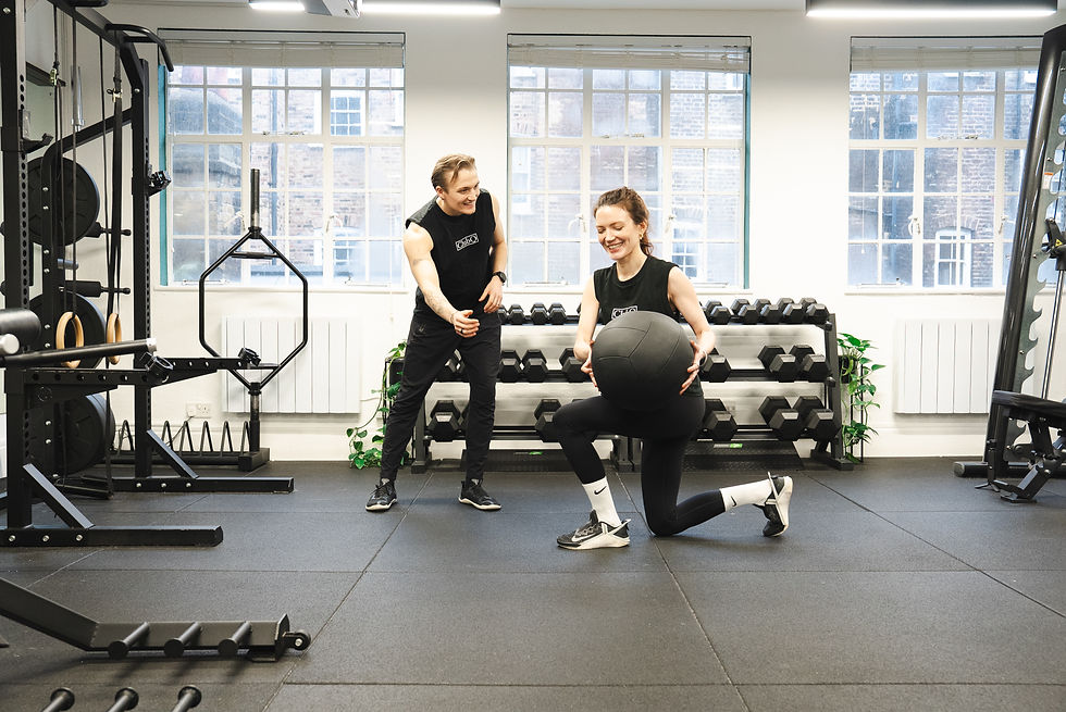 Man coaches a woman doing lunges with a medicine ball. They're in a bright gym with weight racks. Both are wearing black athletic gear, smiling.