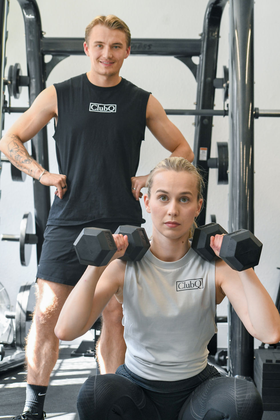 A woman in a grey vest with a ClubQ logo on it is doing squats while a man in a black shirt (with a ClubQ Logo) is standing behind her smiling