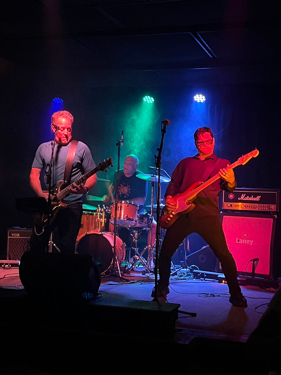 Wide landscape photo of a three-piece rock band performing live on stage with colorful lighting