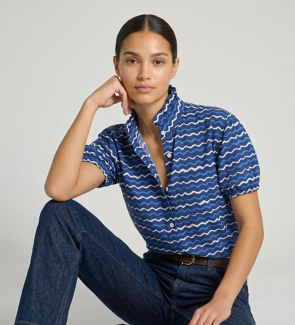 Model seated on the floor wearing a blue zigzag cotton frill‑neck blouse with dark jeans against a light grey backdrop.
