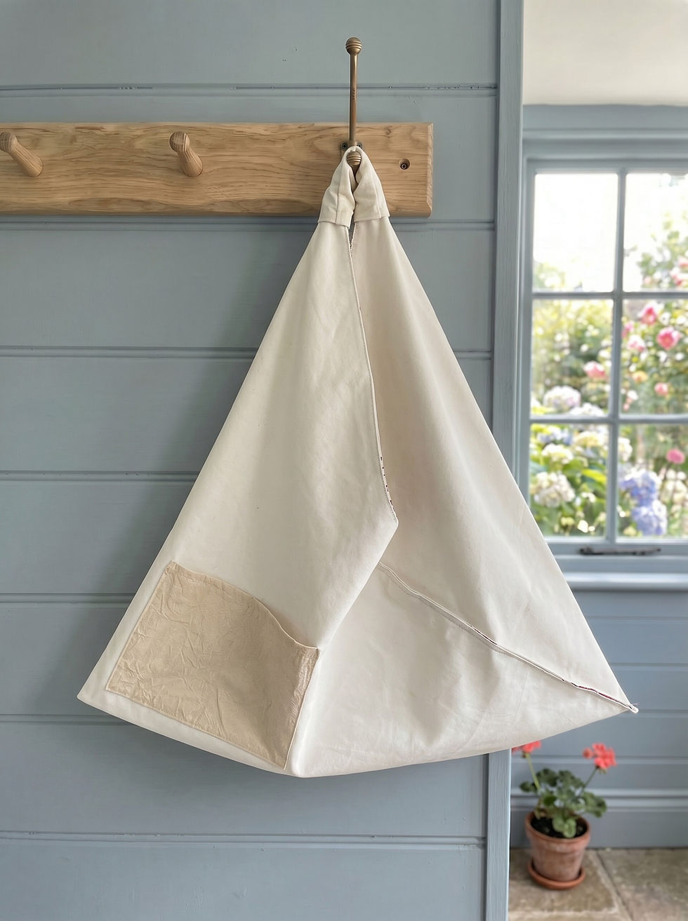 Cream calico‑lined fabric bag hanging from a brass hook on a wooden rack with a garden view in the background