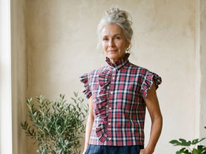 Model with grey up-styled hair wearing a red, blue and white plaid ruffled blouse with navy trousers, standing on a wooden floor near a window and indoor plants.
