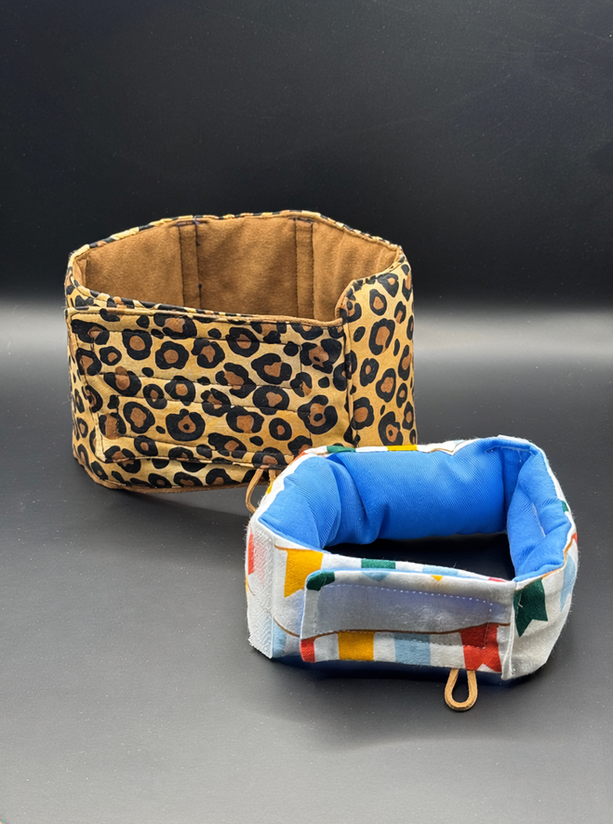 Leopard‑print CaniFeed dog support collar and a smaller geometric FeliFeed cat collar displayed side by side on a dark background. Both are padded and upright, with the dog collar featuring a side loop for attaching an ID tag.