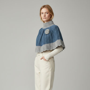 Model wearing a hand‑crocheted blue and grey capelet with ribbed collar and floral detail, styled over a light top and cream trousers.