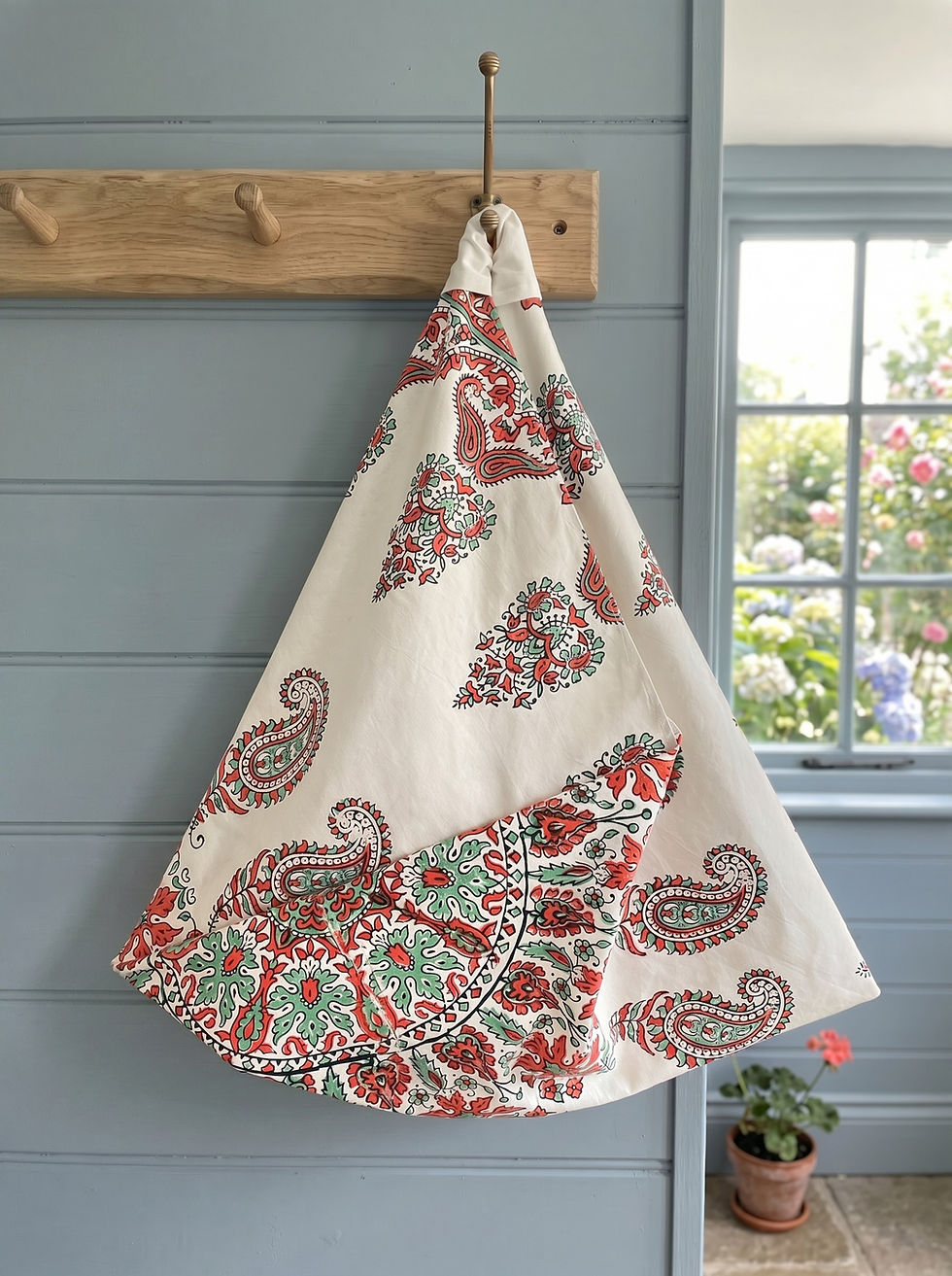 Indian block‑printed Azuma shoulder bag with a red and green paisley pattern hanging on a wooden peg rack by a window.