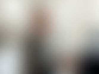 Elderly woman happily plays piano in a bright room with large windows. A vase of flowers is on a table in the background.