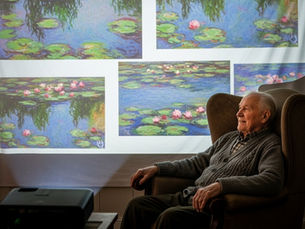 Elderly man seated in armchair, smiling at vivid Monet water lily paintings projected on the wall; calm, contemplative mood.