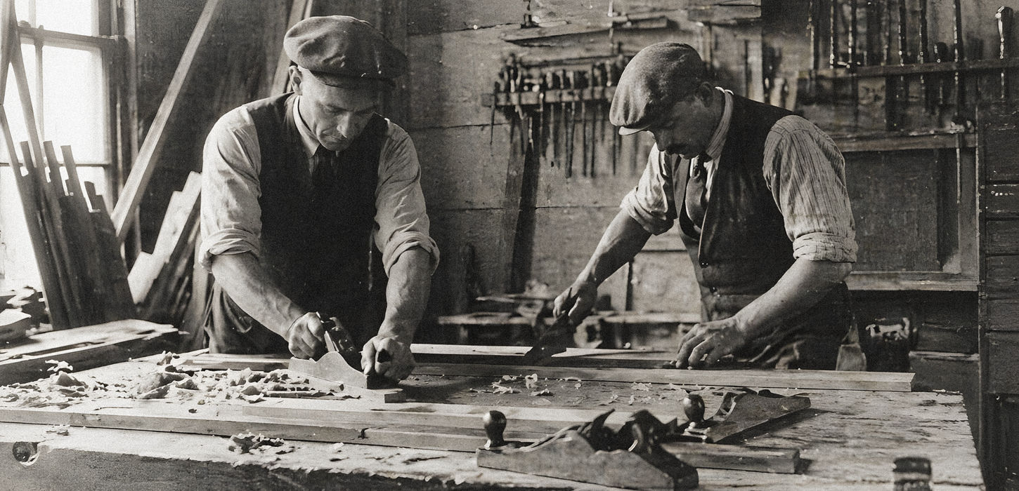 Black and white photograph of two men using hand planes to smooth a large wooden board on a workbench in a traditional woodworking shop, with tools hanging on the wall behind them.