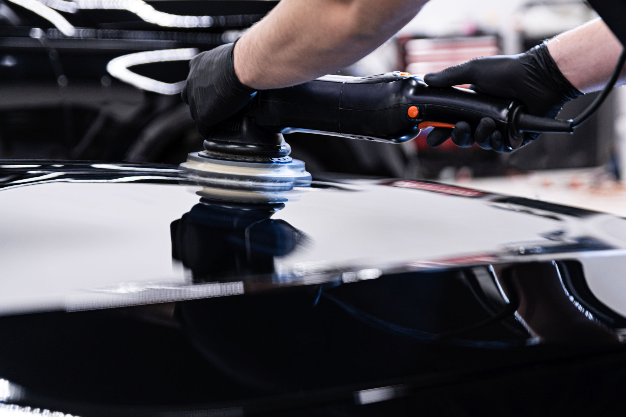 A professional auto detailer wearing black gloves uses a polishing machine on the shiny surface of a black car, highlighting the meticulous care and expertise provided by King's Auto Detailing. The background features a blurred workshop setting, emphasizing a focus on the detailing process.