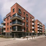 Corner view of a modern residential building clad in red brick with projecting balconies, dark brick base, and white upper levels along a quiet street.