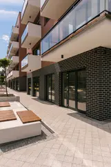 Ground-level detail showing red brick apartments with glass balconies above a dark brick base and storefront-style entrances along a paved sidewalk.