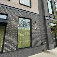 Street-level detail of dark gray thin brick exterior with large storefront windows, wall-mounted lighting, and a stone base along the sidewalk.