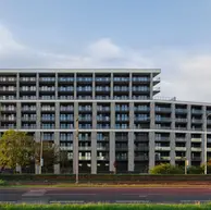 Wide street-level view of a contemporary mid-rise residential building with a long, stepped facade clad in light gray brick and repeating balconies.