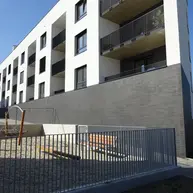 Wide exterior view of a modern apartment building with white upper floors, dark thin brick base, recessed balconies, and a small fenced outdoor seating area.