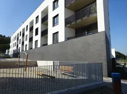Wide exterior view of a modern apartment building with white upper floors, dark thin brick base, recessed balconies, and a small fenced outdoor seating area.