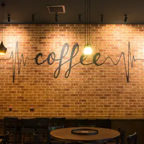 Coffee shop interior with a large exposed thin brick feature wall in blended red and earthy brown tones, decorated with the word “coffee” in black script and illuminated by hanging pendant lights.