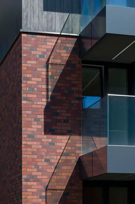 Close-up of Gothic Smile and Old Factory thin brick in king size, showing warm red clay tones with subtle dark charcoal accents. The brick’s slightly varied surface texture and elongated king-size proportions create a refined, linear look beneath modern glass balconies and dark cladding.