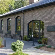 Side elevation of Oficyna Restaurant showcasing Aztec Ghost thin brick in soft gray-beige hues, arched window openings, black-framed glazing, and outdoor seating along a paved walkway.