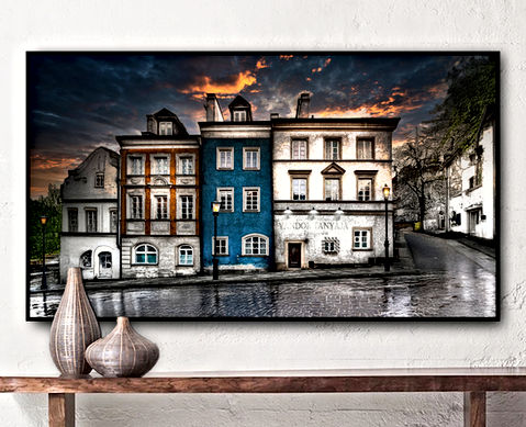 The artwork captures a moody, romantic European street scene at dusk. A row of historic, charming buildings lines a cobbled street glistening with recent rain. The architecture features a blend of baroque and rustic elements with textured facades—white, burnt orange, and a striking blue centerpiece. The words "Vándor Tanyája Szálloda" are subtly inscribed on the wall of the rightmost building, hinting at an old-world inn or hotel, loosely translating from Hungarian to "Wanderer's Lodge Hotel." A dramatic sky smolders with deep blues and fiery orange clouds as vintage-style street lamps cast a soft glow. In the foreground, earthy-toned vases sit on a minimalist wooden table, suggesting the image is printed on canvas and hung as art in a cozy, modern space.