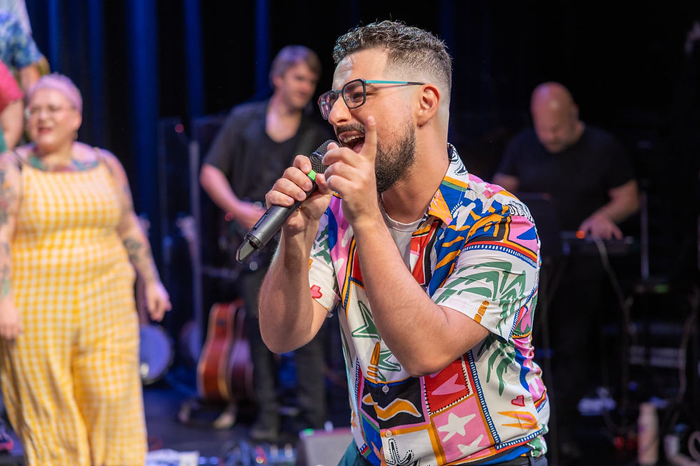 A soloist from YVR Pop Choir at Chorus Studio performs on stage in Vancouver wearing a colorful patterned shirt, singing expressively into a microphone with the live band playing behind him during a high-energy concert.