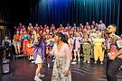 A vibrant moment on stage at Chorus Studio: Rebecca Lam passionately directs a choir of over 70 diverse singers, all raising their arms and belting out in full voice. Two soloists perform joyfully at the front, and a band member stands to the right holding a mic. The stage is full of colour, energy, and community spirit.