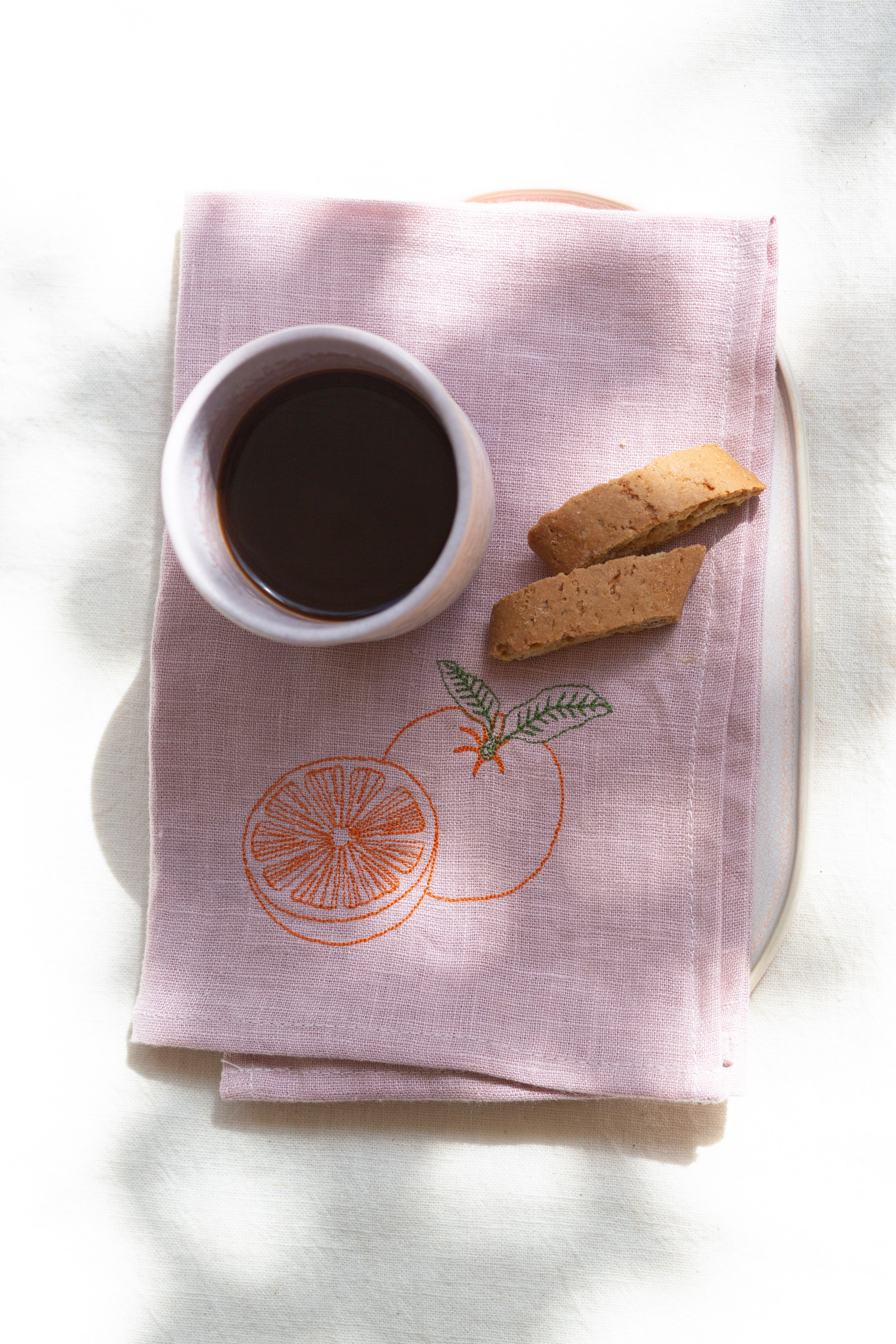 Rosa Leinenserviette mit Orangen-Stickerei auf einem ovalen Teller mit einem Becher Kaffee und zwei Keksen