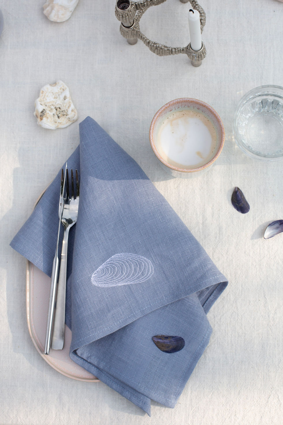 Hellblaue Leinenserviette mit Miesmuschel-Stickerei auf einem ovalen Teller mit silbernem Besteck dekoriert mit Muscheln