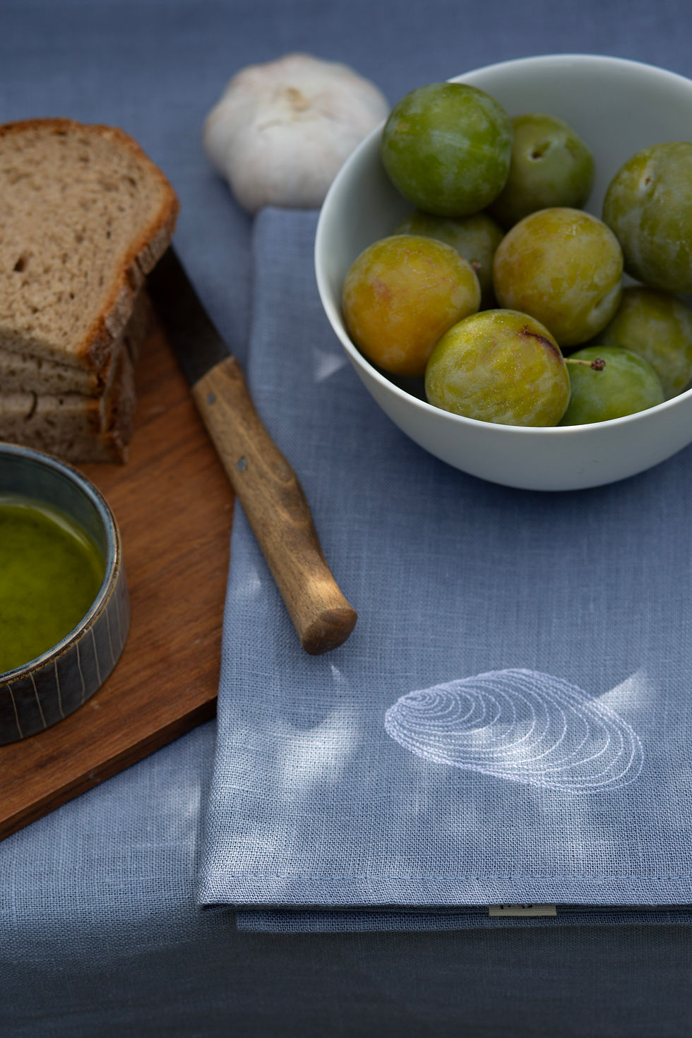 Miniaturbild: Hellblaue Leinenserviette mit Miesmuschel-Stickerei unter einer Schale mit gelben Pflaumen neben frischem Brot