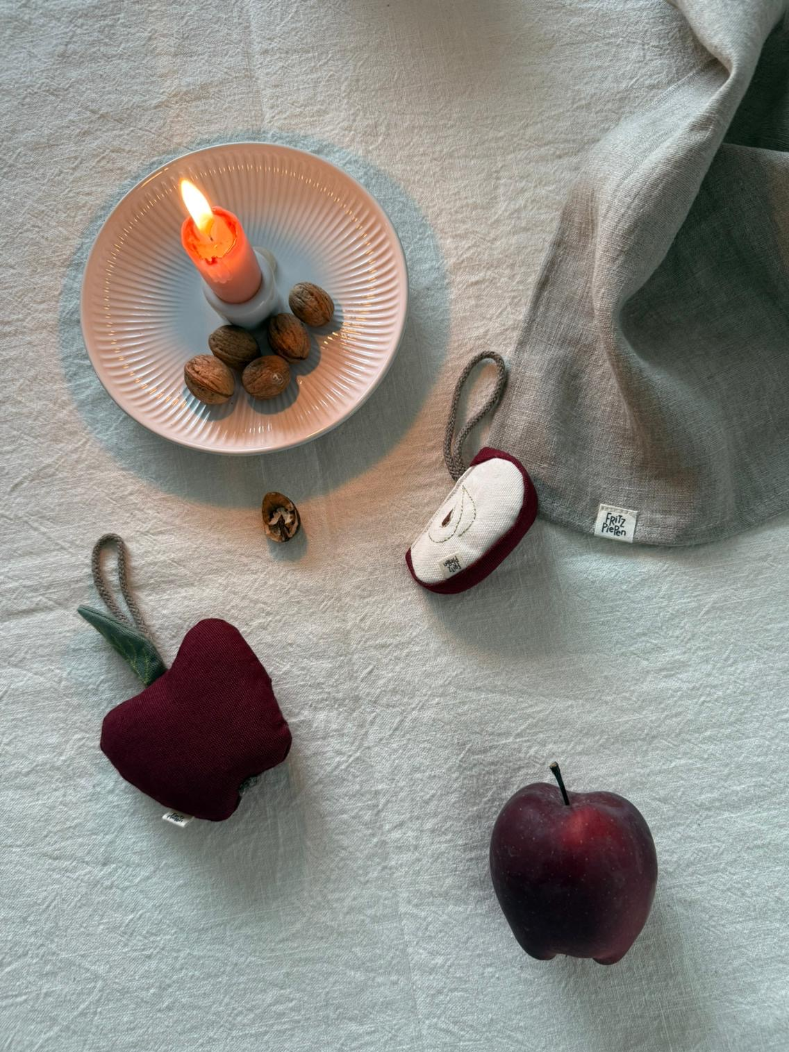 handgefertigter Apfel aus Stoff mit dezenten Stickdetails, liegend auf heller Tischdecke mit Kerze dekoriert