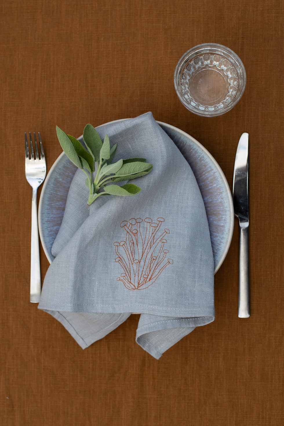 Hellgraue Leinenserviette mit Enoki-Pilzstickerei auf einem Teller dekoriert mit Salbei auf goldbrauner Tischdecke