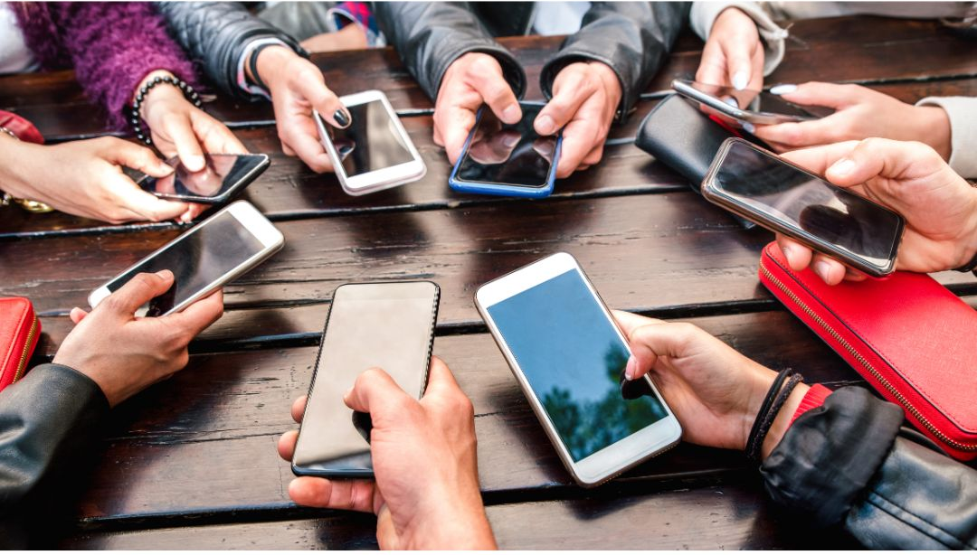 Several people hold smartphones around a wooden table, focused on their screens in a casual social setting.