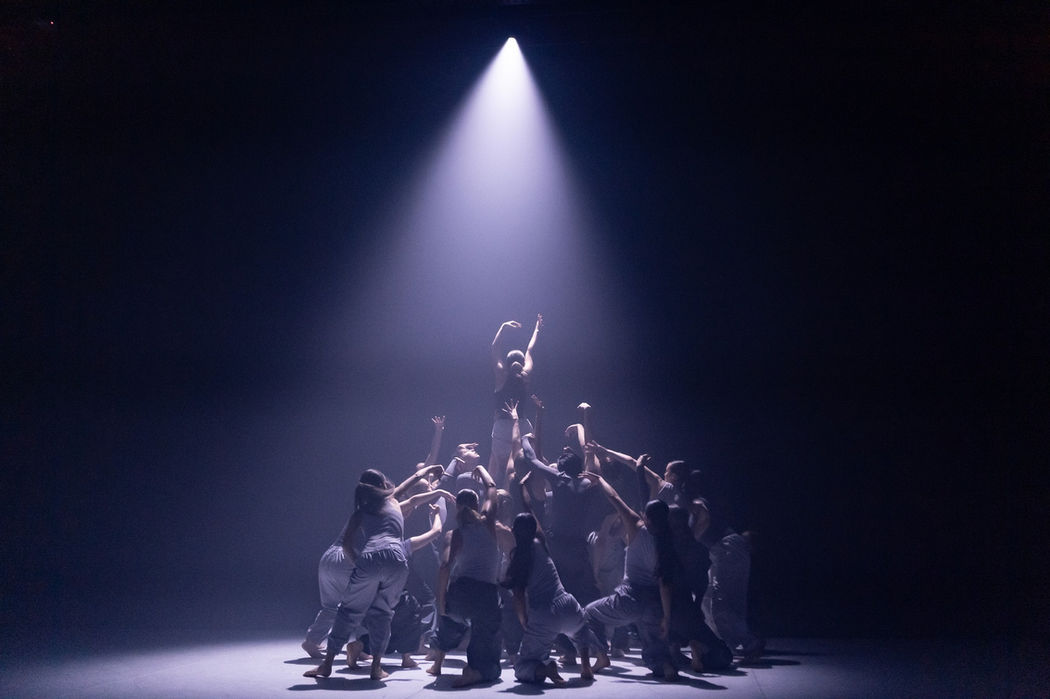 A large group of performers lifting one into the air. They are lit by one cold white downlight.