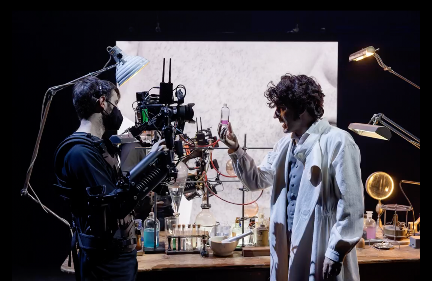 A man in a white lab coat holding a small vial filled with a pink liquid, a camera operator shoots directly on, both are standing in front of a cluttered lab table and a square digital screen displaying a section of the cameras live feed.