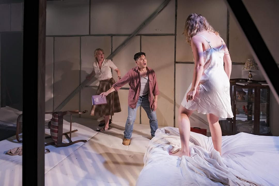 A woman in a silk dress stands menacingly on a bed. A man in a red shirt and jeans, holding a Bible, shields a woman in a white shirt and plaid skirt. They are lit in warm white.