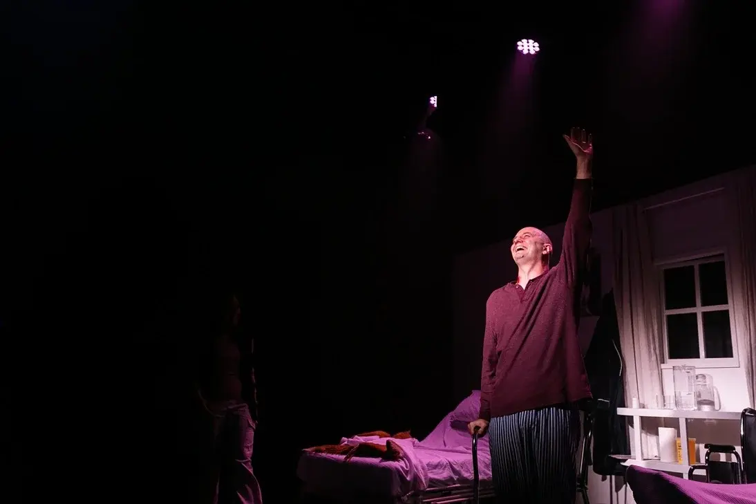 A man in a maroon jumper stands with his arm up, in the middle of hospice room. He is lit in white, the room in pink.