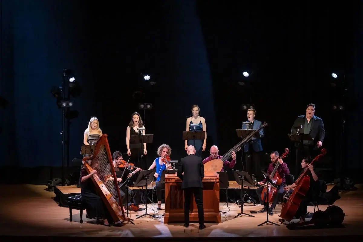 A wide shot of City Recital Hall. On stage, a conductor leads a six-piece orchestra and on raised platforms behind them, five opera singers. They are lit in cold white.