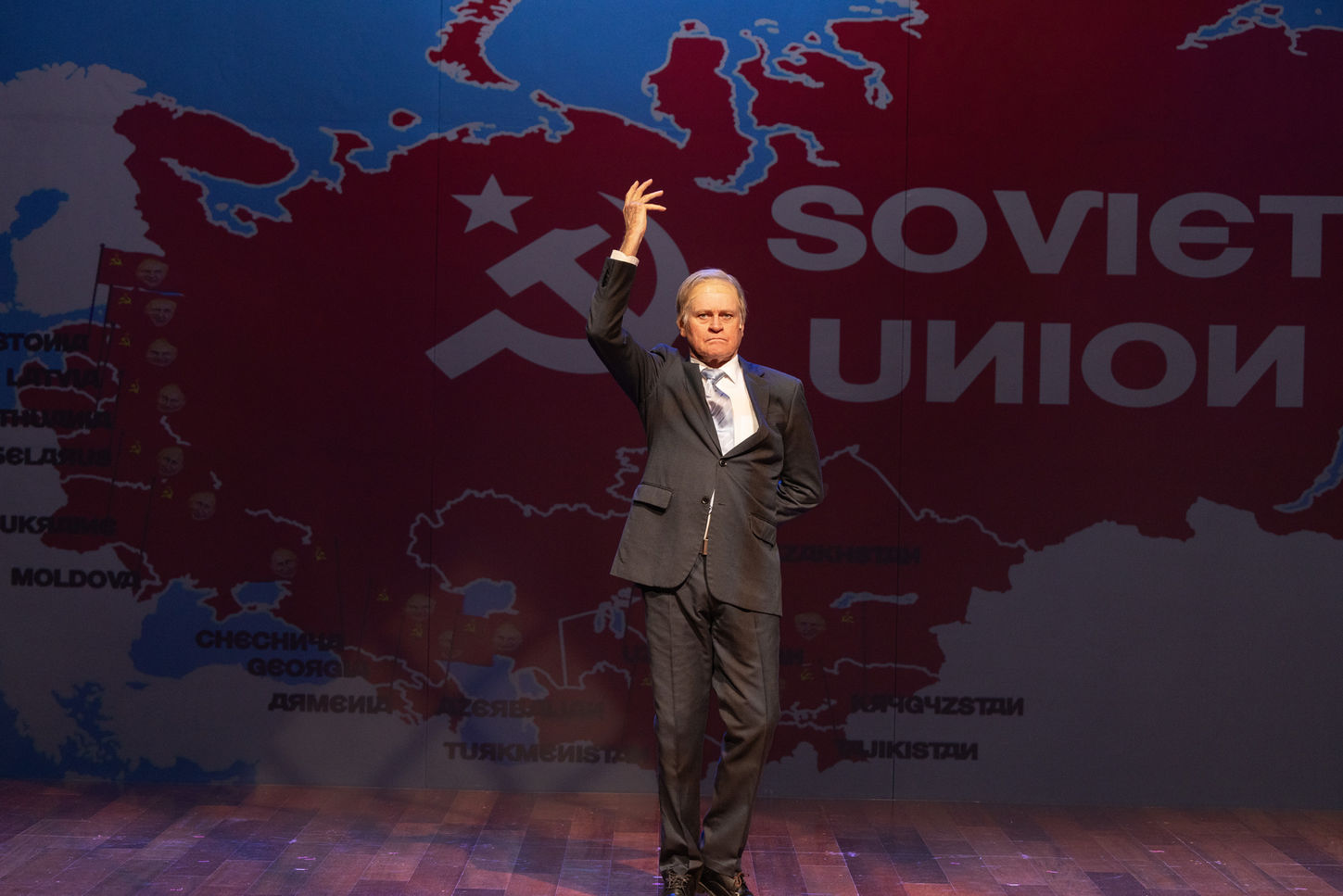 A man in a grey suit and wig, standing with his hand in the air. A large projected image of a map of the Soviet Union and the surrounding countries being taken over is visible in the background. He is lit in warm white.