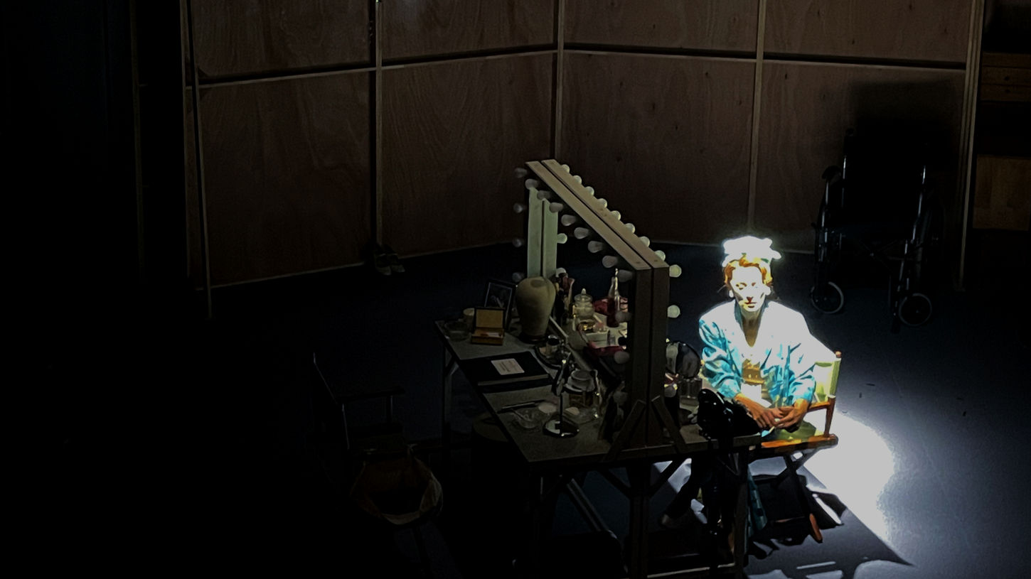A woman dressed in blue, seated in a director's chair at a makeup table. She is lit in white. Behind, projected on the wall, a large live-feed image of her face in black and white.