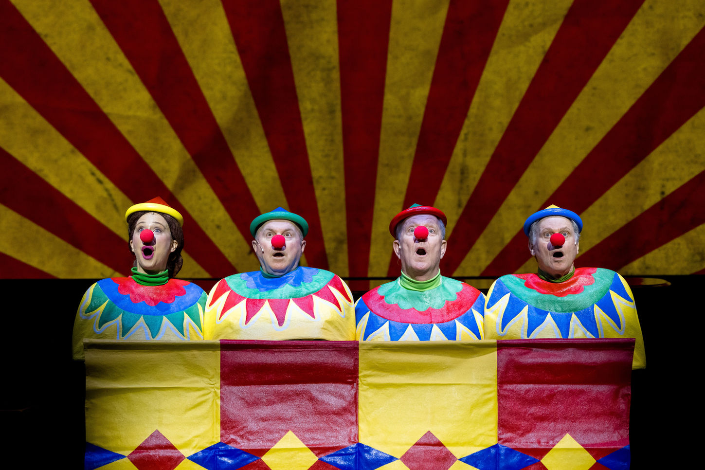Four performers dressed as “Laughing Clowns” carnival game clowns, wearing colourful hats and red noses, seated behind a red and yellow bannister. In the background, a large projected image of a red and yellow striped circus tent is visible. They are lit in cool white.