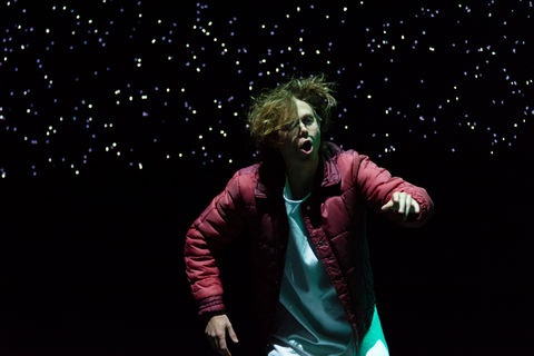 A man in a red puffer jacket, pointing to something off-camera. A long, thin digital screen displaying a star field is visible in the background. He is lit in cold white. 