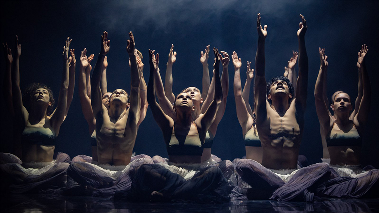 Sixteen performers, seated cross-legged on the ground, wearing white ruffled pants, the females in black tops. They are in five columns, with two rows on the outside columns and four on the inside. They are lit in cool white. 