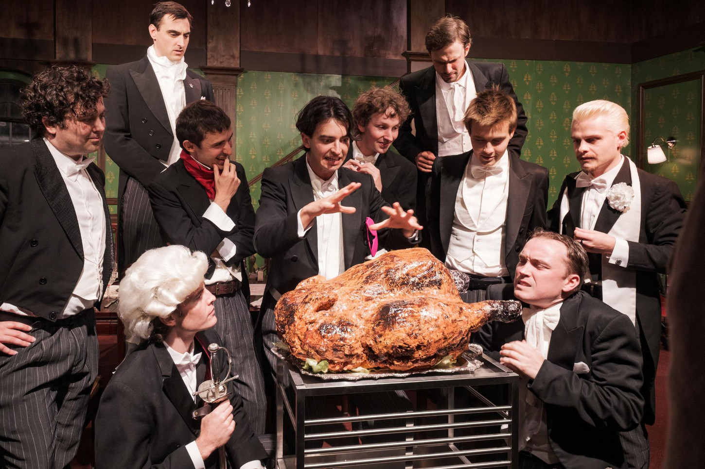 Ten men dressed in full tails stand around a large cooked chicken in amazement. They are lit in warm white.