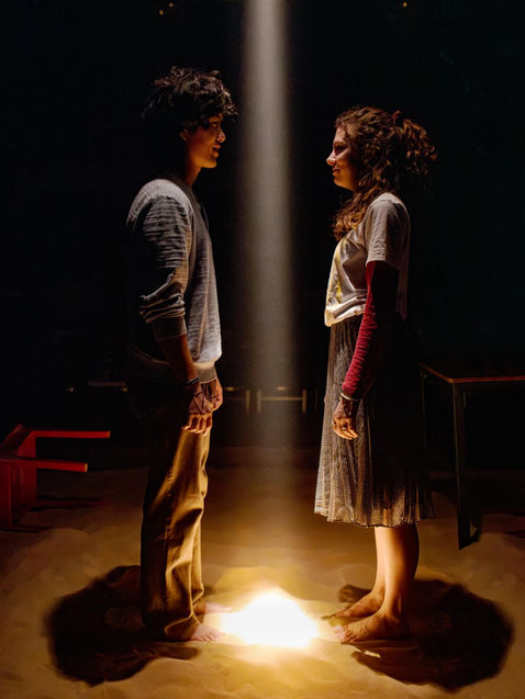 A man in a grey shirt and a woman in a white shirt and grey skirt, stand in sand looking at each other, split by a single warm light shooting directly downward onto the sand. They are both lit by this light.