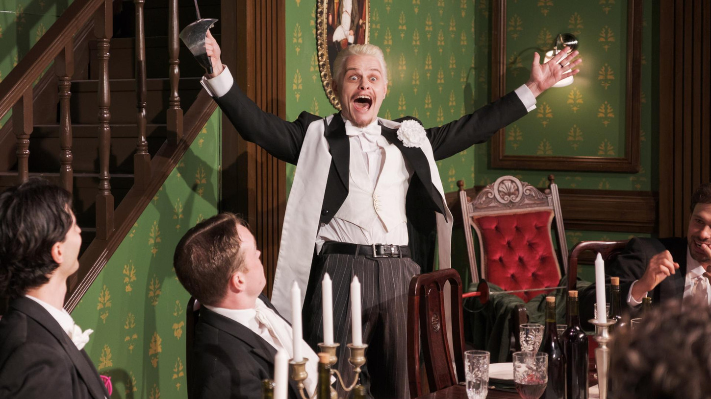 A man dressed in full tails stands arms outstretched at the head of an elegant dining table. Other men also in full tails sit around it. They are lit in warm white.