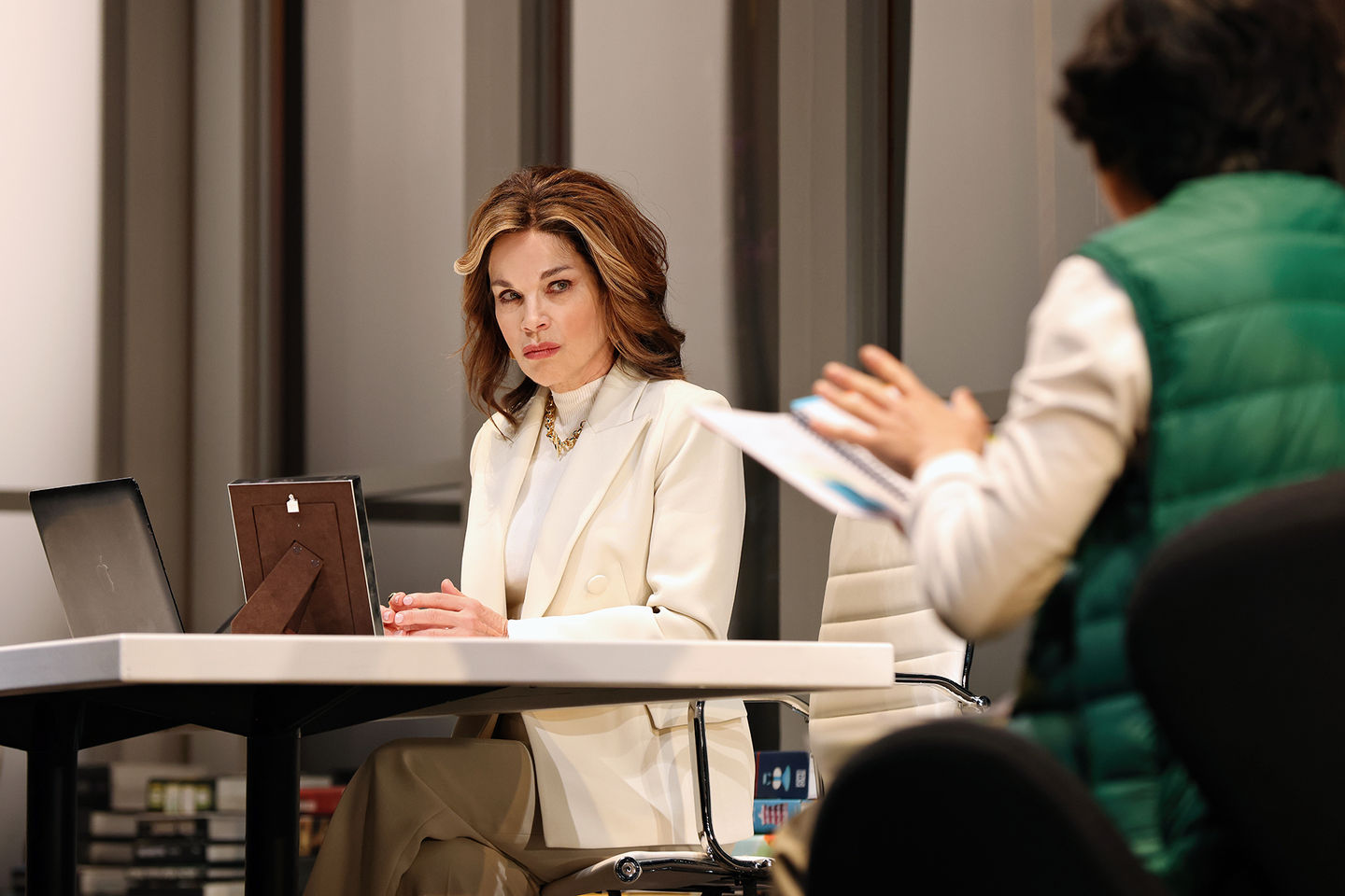 A woman in a white blazer seated behind a desk looking at a man in a green sleeveless puffer jacket holding a note book, with his back to camera.