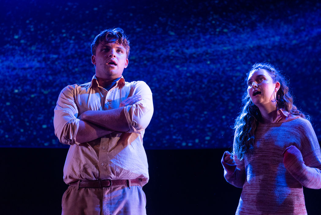 A man, centred, arms folded, a woman to his right, both looking out. A large, curved projected image of the night sky is visible in the background. Both lit in purple.